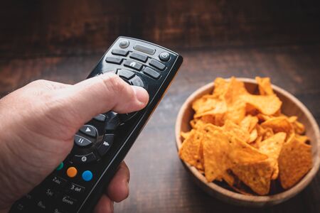Chips in a wooden bowl next to the TV remote control against a burnt wooden background. Top view....