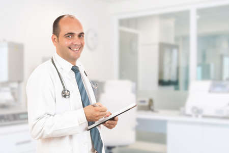 Male doctor with stethoscope holding a notebook. Smiling male doctor filling out medical form on clipboard while standing straight in hospitalの写真素材
