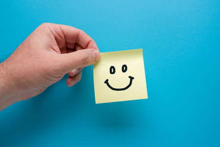 Hand holding yellow sticky note paper or post-it with smiley icon on blue backgroundの写真素材