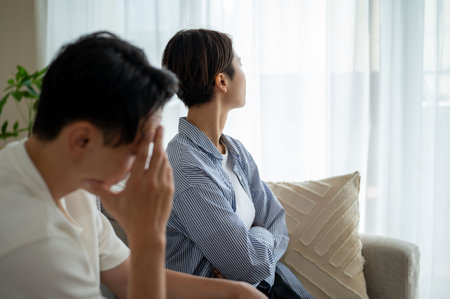 Depressed Asian couple sitting on sofa and looking at each other at homeの写真素材