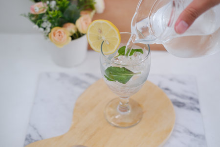 Pouring water into glass with mint and lemon on white marble table