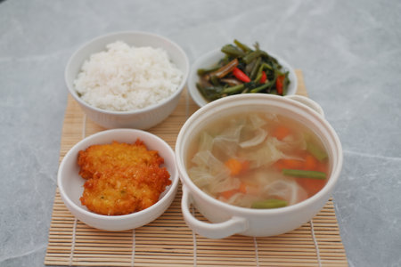 The set includes a bowl of clear vegetable soup filled with cabbage, carrots, and green beans, served in a white bowl with handlesの写真素材
