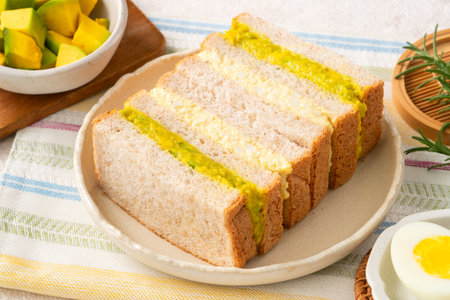 Delicious soft sandwiches filled with mashed creamy avocado and egg salad on a light breakfast table.の写真素材