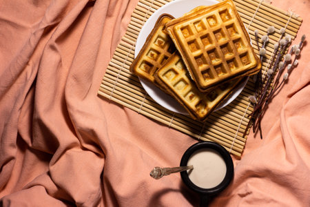 top view of belgian waffles on white plate and bamboo mat with willow twig and earthenware cup of ryazanka with spoon on pink backgroundの写真素材