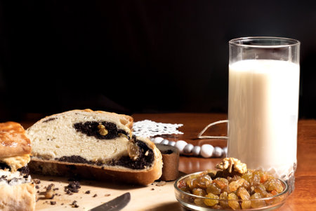 ruddy slices of poppy seed roll with crumbs on a bamboo wooden cutting board with a knife and a glass of milk on a dark wooden table with a bowl of raisins and a white Christmas tree toy with a black backgroundの写真素材