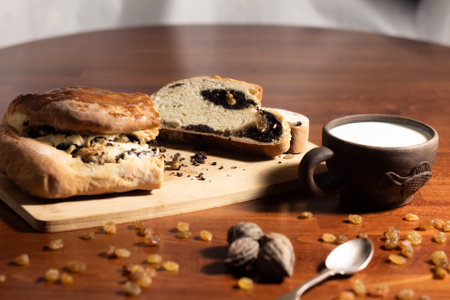 ruddy slices of poppy seed roll with crumbs on a bamboo wooden cutting board with a clay cup with milk on a dark wooden table with scattered raisins and nuts and a blurred backgroundの写真素材