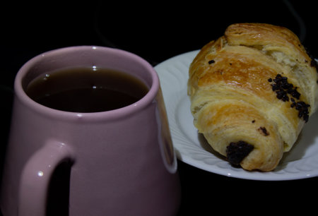 a cup of tea and a croissant with chocolate on a white plate on a black background high angle view