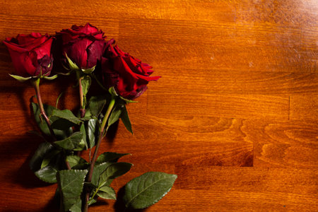 Red roses with lush green leaves are arranged neatly on a warm wooden table. The gentle light highlights the deep hues of the petals and the texture of the wood.の写真素材