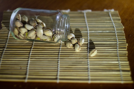 A glass jar filled with pistachio nuts lies open on a bamboo mat, with some nuts spilled out, creating a natural and rustic atmosphere perfect for culinary, health, or snack-related themes.の写真素材