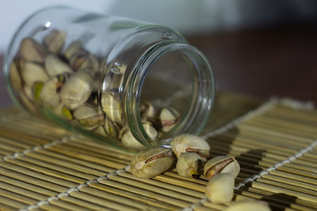 Pistachios scattered from a glass jar on a bamboo mat, creating an earthy and natural setup with a...