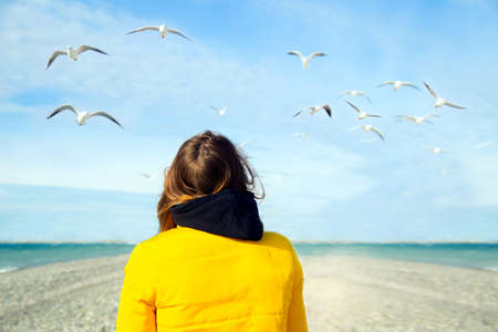 back view girl wearing yellow jacket on the shore with seagulls at sky. girl traveller relaxing on the beach and feeds the birds.の写真素材