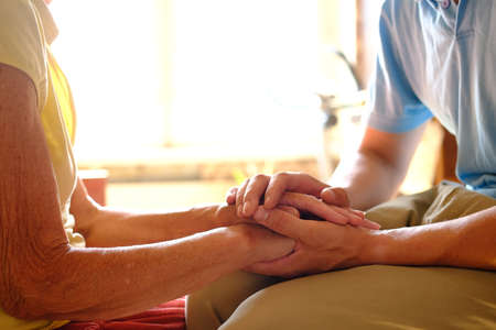 Worried caucasian man gently stroking hand of his sick old mother showing care or love. Son comforting wrinkled arm of elderly mom sitting at bed. Guy giving support to his old parent slow motion.の写真素材