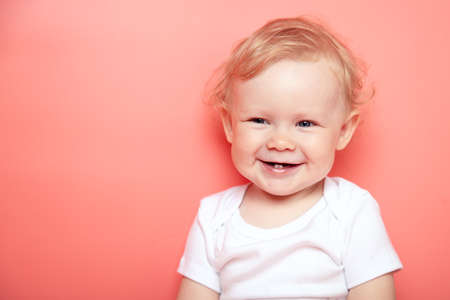 portrait caucasian curly blond smiling baby girl with two teethon pink background wearing in white...