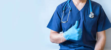 Cropped image of handsome male surgeon doctor with stethoscope in blue uniform with statoscope holding thumb up in clinic on blue background. banner with copy spaceの写真素材