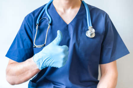 Cropped image of handsome male surgeon doctor with stethoscope in blue uniform with statoscope holding thumb up in clinic on blue background. banner with copy spaceの写真素材