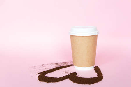 eco disposable paper glass with ground coffee shape of heart on soft pink background.