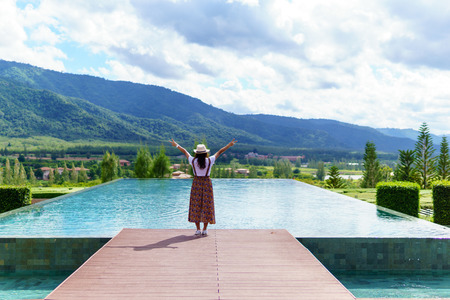 rear view of young asian female standing near the pool and put her hand up for amazing panorama...