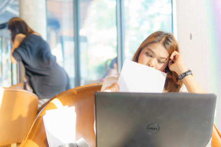 beautiful asian young business woman feeling tired and sit near transparent window and hold business document with laptop for her job at cafe or coffee shop. business concept.の写真素材