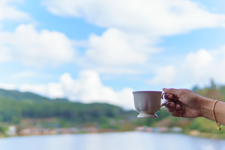 asian female hand  holding  white coffee  or tea cup with cloudy blue sky, lake and mountain background.の写真素材
