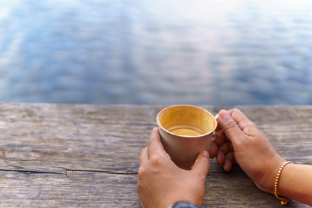 asian female hand  holding  white coffee  or tea cup  on the wooden plank near pond or lake.の写真素材