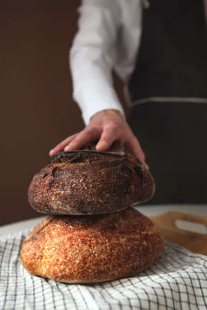 Man wearing apron holding hand on the top of two warm white and brown loafs of homemade bread on the brown backgroundの写真素材