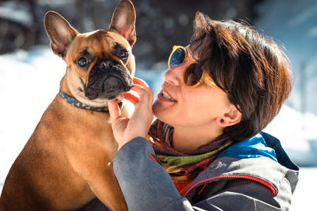 Woman wearing sunglasses holding on hands French buldog and playing with it on the white snowy background. Dog looking in cameraの写真素材
