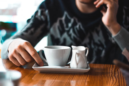 Man talking over the phone in a cafe with a coffee cup and cream milk pot on the table. Adult hand and winter sweaterの写真素材