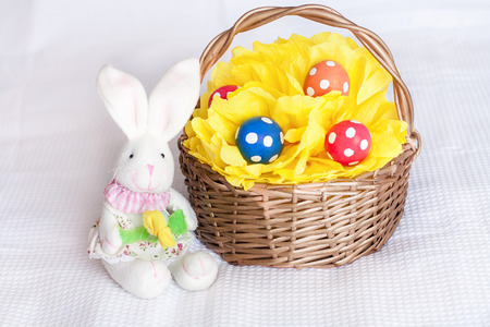 Easter eggs on yellow pompom in wicker basket and toy rabbit on checkered white backgroundの写真素材