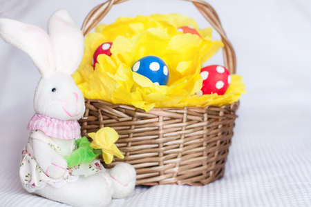 Easter eggs on yellow pompom in wicker basket and toy rabbit on checkered white backgroundの写真素材