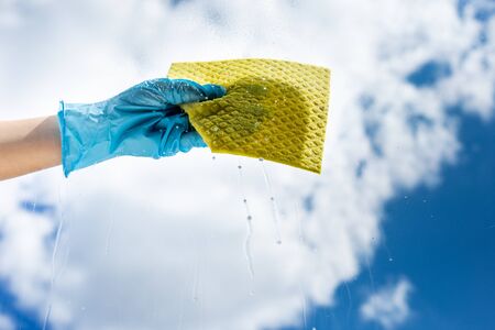 Human hand holding napkin for cleaning window on grey background. A female hands in bright blue gloves washes the windows. The concept of cleanlinessの写真素材