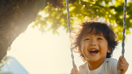 Cheerful Asian kid boy laughing on a swing on a warm sunny day on a playground. Concept of carefree play, happy childhood, summer fun, and outdoor activities.の素材