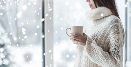 Caucasian woman in a cozy white sweater holding a steaming mug by a window. Concept of warmth winter...