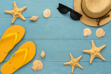 Beach essentials flat lay featuring yellow flip-flops, straw hat, sunglasses, and seashells on a blue wooden background. concept of summer vacation, ocean vibes, relaxation, travel. Copy space.の写真素材