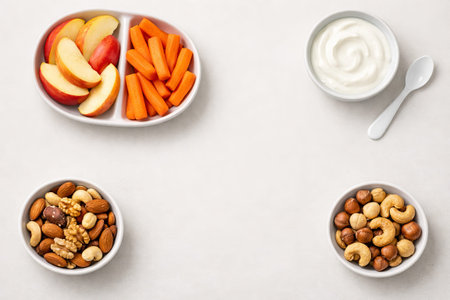 Healthy snack options with fresh apple slices, carrot sticks, mixed nuts, and yogurt in bowls. Clean...