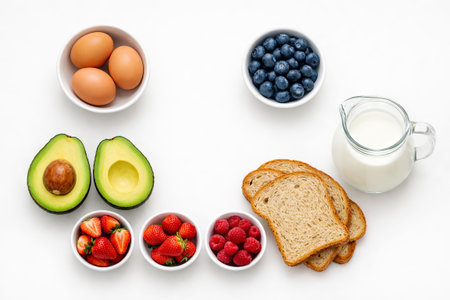 Healthy breakfast food items arranged on white background, including eggs, avocado, berries, bread,...