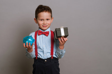 a smart boy 5-7 years old in a business image on a gray background holds a pig piggy bank in his handsの写真素材
