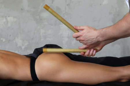 Massage with bamboo sticks for a young girl. Masseur makes anti-cellulite massage with bamboo sticks on thighs, legs and buttocks in a beauty salon. Cellulite and Overweight Treatment.の写真素材