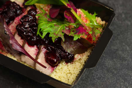 Healthy box with turkey with cherry, greens and couscous in eco paper black takeaway container on dark background. Restaurant food delivery concept.の写真素材