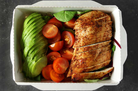 Healthy lunch box with chicken fillet, avocado, tomatoes and lettuce on dark background