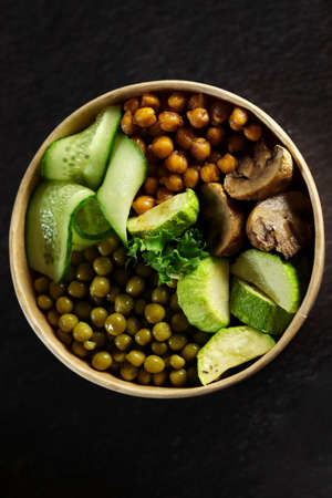 Healthy lunch bowl with chickpeas, mushrooms, cucumber, green peas and hummus on dark background,...
