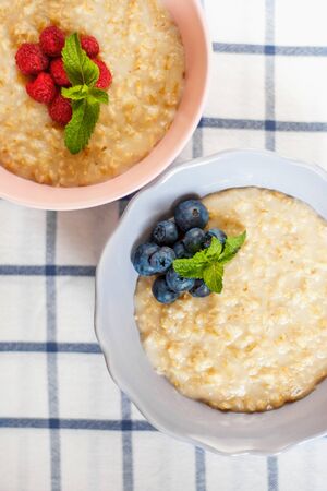 Healthy homemade oatmeal porridge with berries for breakfast in ceramic bowl on a checkered tablecloth. Morning oatmeal porridge with fresh raspberries and blueberries. Vertical photo. Close upの写真素材