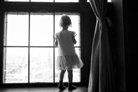 Child is standing and looking out the window. Stay at home and quarantine concept. Sadness of the toddler.の写真素材