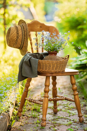 Vintage wooden chair standing in the garden with wicker basket, ceramic vase and bouquet of forget-me-not's on itの写真素材