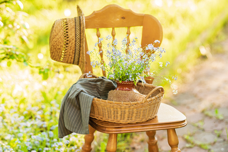 Vintage wooden chair standing in the garden with wicker basket, ceramic vase and bouquet of forget-me-not's on itの写真素材