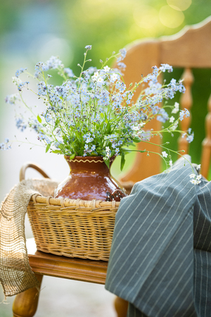 Vintage wooden chair standing in the garden with wicker basket, ceramic vase and bouquet of forget-me-not's on it. Close upの写真素材