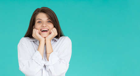 Indoor portrait of young attractive European girl isolated on blue background looking straight at camera propping up her chin with fists feeling great joy, smiling and laughing at jokeの写真素材