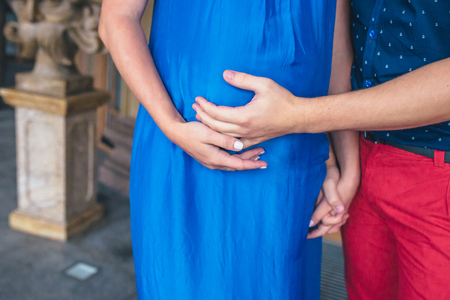 The future father supports his wife's pregnant belly. Future mom in a blue dress with white manicure, dad in a red pants and dark blue shirt.の写真素材