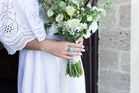 The ring with blue stone on the bride's finger. The bride in white wedding dress holds a wedding bouquet with white roses, poppies and greenery decorated with long silk ribbonの写真素材
