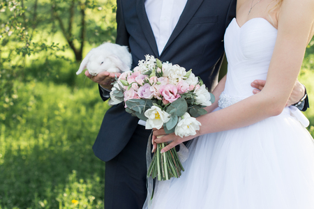 Wedding day. Bride holding in her hands a delicate wedding bouquet with white and pink tulips and pink small roses. Groom holding a white cute rabbit.の写真素材