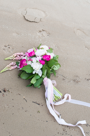 Delicate bridal bouquet with white and pink peony and greenery with long ribbon lying on wet sand on the bank of riverの写真素材